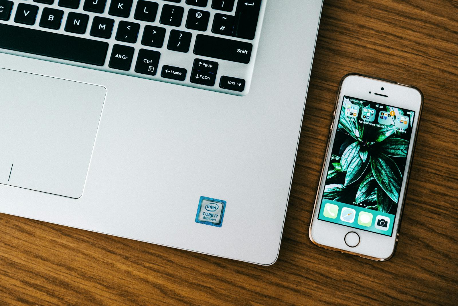 iPhone with home screen visible beside a laptop keyboard on a wooden desk — photo by Fotios Photos on Pexels