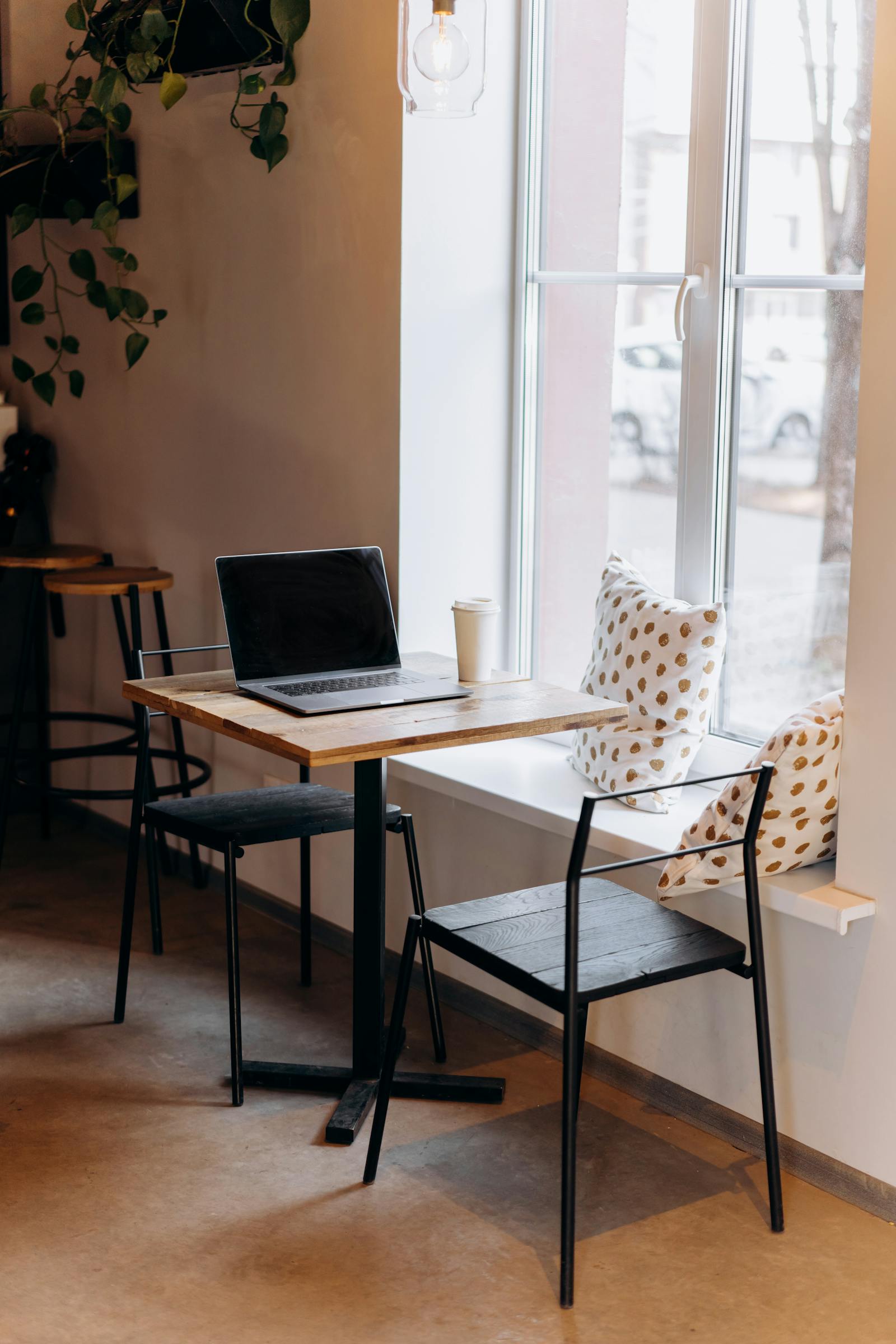 Laptop on a wooden cafe table by a window with hanging plants — photo by Mikhail Nilov on Pexels