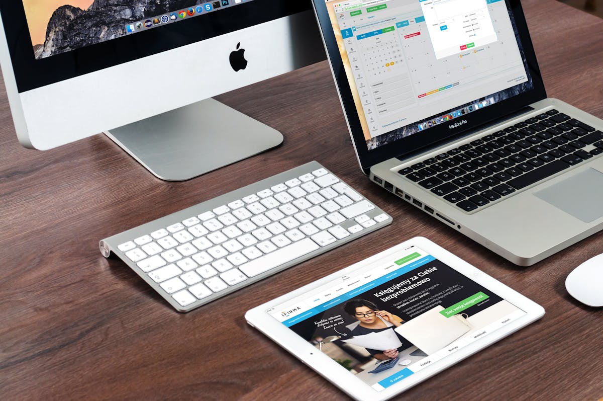 An iMac, a MacBook Pro, and an iPad arranged together on a wooden desk with a Magic Keyboard and Magic Mouse — typical multi-Apple-device workspace where Universal Control matters (photo: Pixabay / Pexels)
